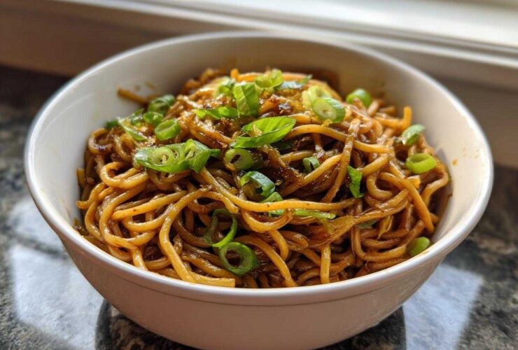 A close-up of rich, saucy garlic noodles topped generously with bright green sliced scallions in a white bowl.