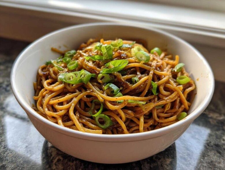 A close-up of rich, saucy garlic noodles topped generously with bright green sliced scallions in a white bowl.