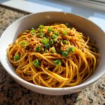 Close-up of savory, saucy garlic noodles topped with chopped green onions and crispy bits in a white bowl.