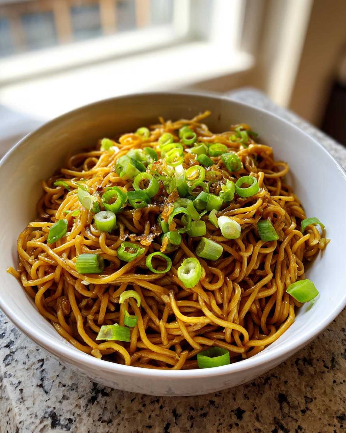 A close-up of a white bowl filled with savory, brown garlic noodles, generously topped with bright green chopped scallions.
