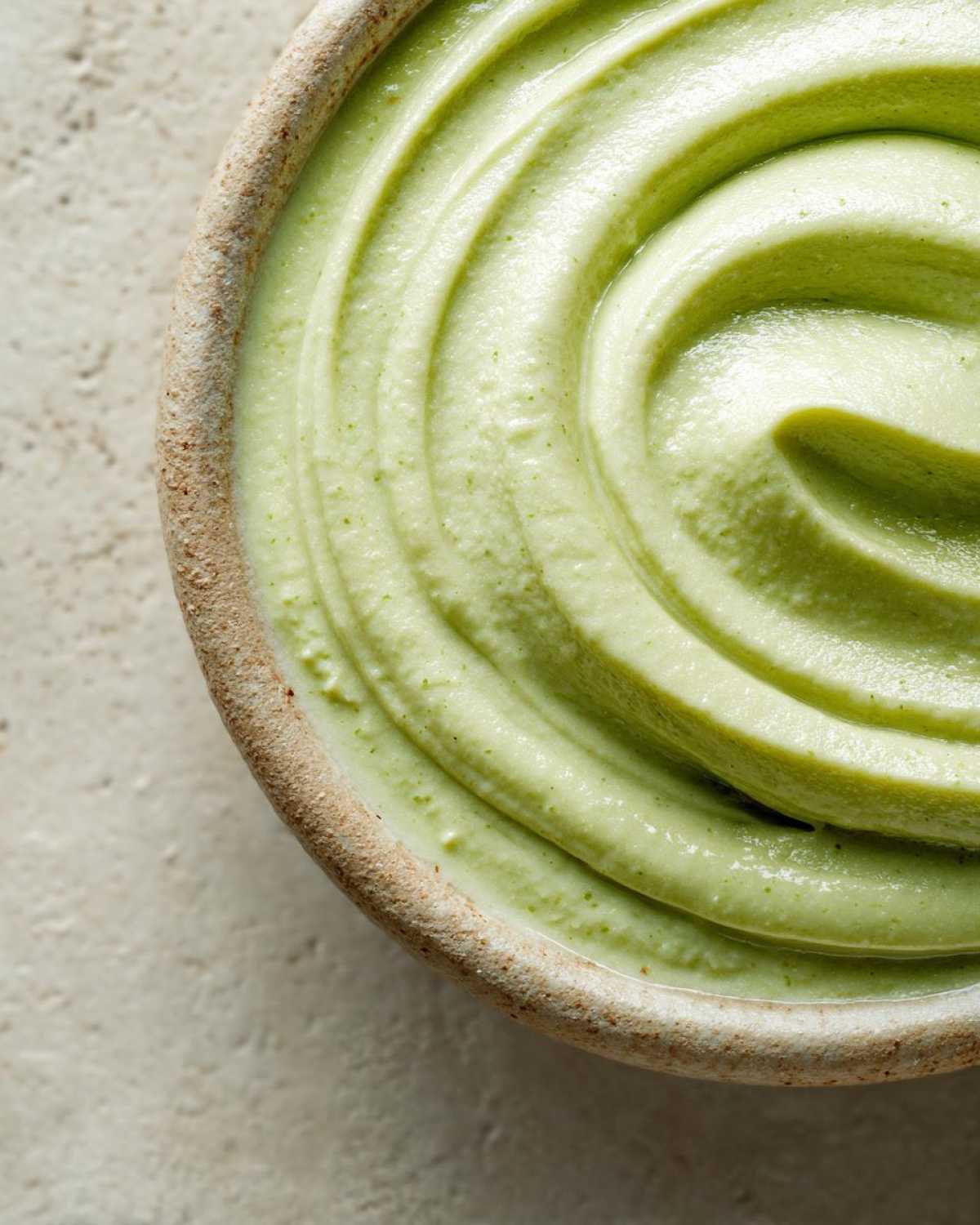 Close-up overhead shot showing the velvety, light green texture of blended celery soup swirled in a rustic bowl.