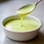 Close-up of a spoonful of bright green, velvety celery soup dripping back into a white bowl.