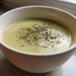 A close-up of a white bowl filled with creamy, velvety leek soup, generously topped with cracked black pepper.