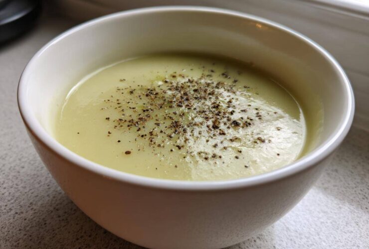 A close-up of a white bowl filled with creamy, velvety leek soup, generously topped with cracked black pepper.