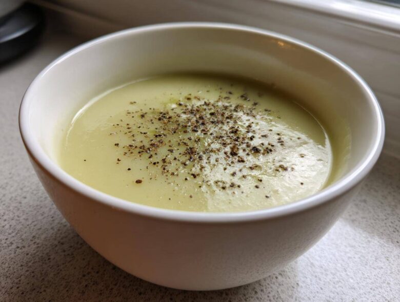 A close-up of a white bowl filled with creamy, velvety leek soup, generously topped with cracked black pepper.