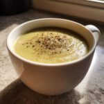 Close-up of a creamy, pale green leek soup served in a white mug, generously topped with freshly cracked black pepper.