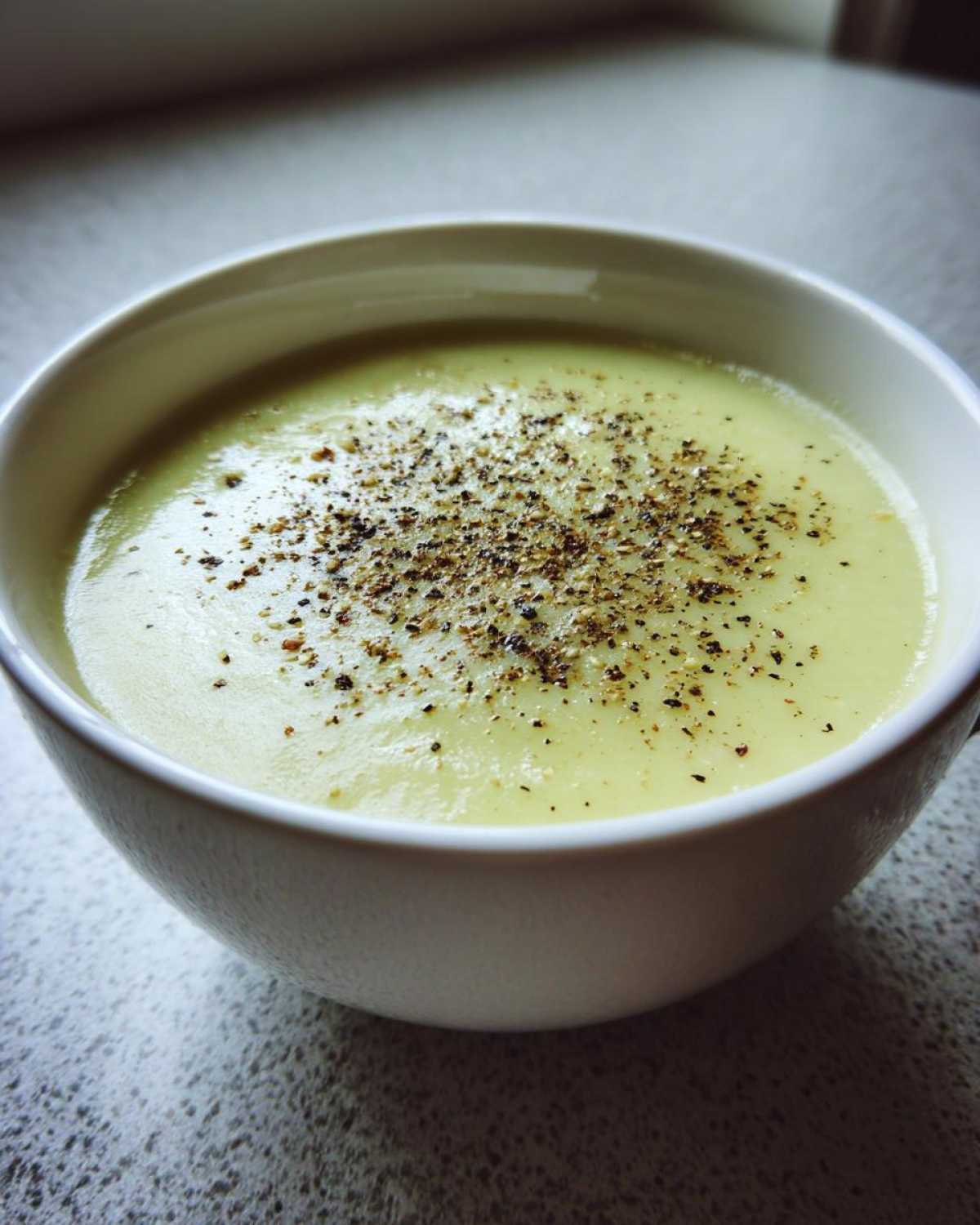Close-up of a bowl of smooth, pale green leek soup generously topped with freshly cracked black pepper.