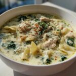A close-up of a white bowl filled with creamy white chicken lasagna soup, featuring noodles, shredded chicken, and spinach.