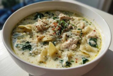 A close-up of a white bowl filled with creamy white chicken lasagna soup, featuring noodles, shredded chicken, and spinach.