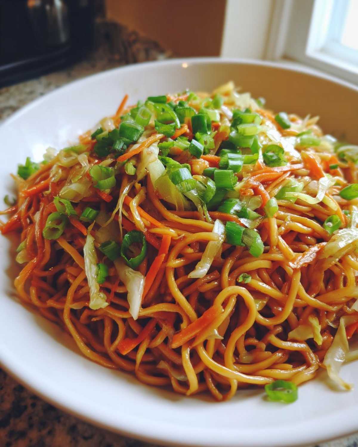 A close-up view of a white bowl filled with savory fried noodles, shredded carrots, cabbage, and topped generously with fresh green onions.