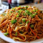 A close-up shot of a generous serving of glossy, brown fried noodles mixed with shredded carrots and topped with fresh green onions.