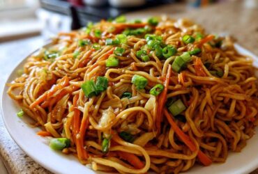 A close-up shot of a generous serving of glossy, brown fried noodles mixed with shredded carrots and topped with fresh green onions.