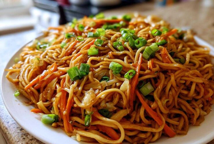 A close-up shot of a generous serving of glossy, brown fried noodles mixed with shredded carrots and topped with fresh green onions.