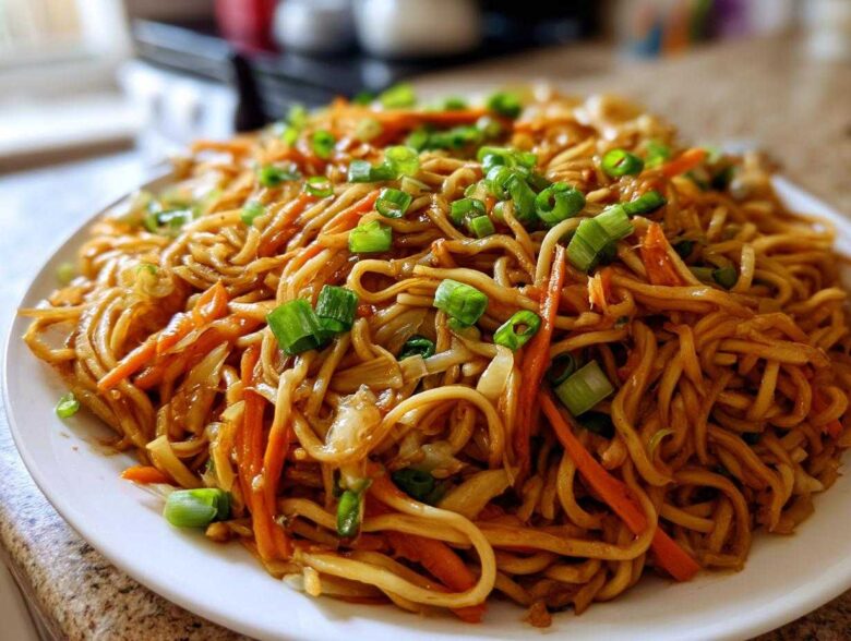 A close-up shot of a generous serving of glossy, brown fried noodles mixed with shredded carrots and topped with fresh green onions.