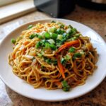 A close-up plate of savory fried noodles mixed with carrots and onions, topped generously with fresh green onions.