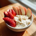 Close-up of a white bowl containing creamy fruit dip surrounded by sliced strawberries and apple wedges.