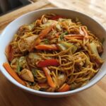 A close-up of a generous serving of homemade chicken chow mein recipe, featuring noodles, chicken pieces, carrots, and cabbage.