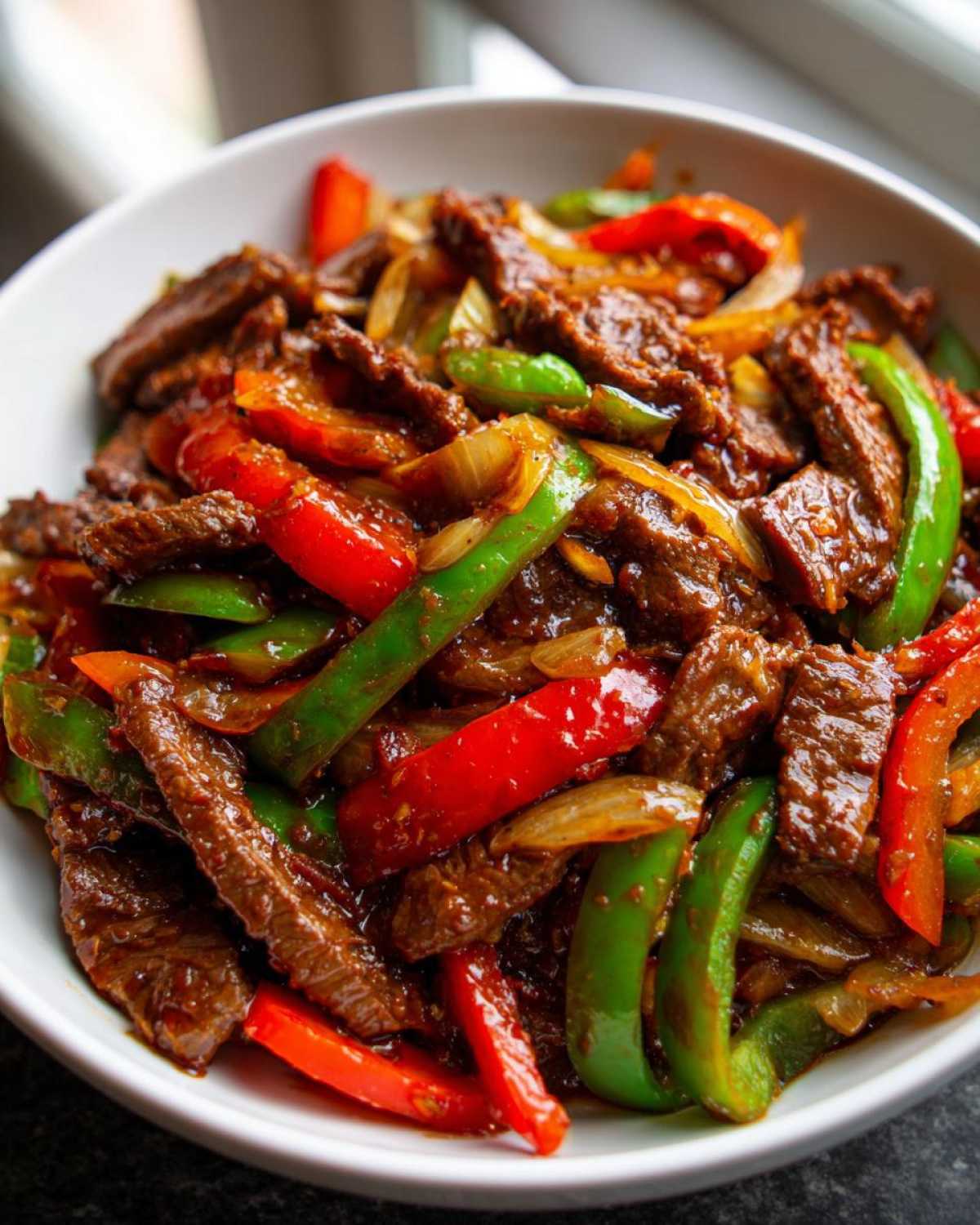 Close-up of savory pepper steak strips mixed with vibrant red and green bell peppers and onions in a white bowl.