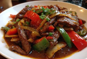 A close-up of a white plate filled with saucy pepper steak featuring strips of beef, red and green bell peppers, and onions.