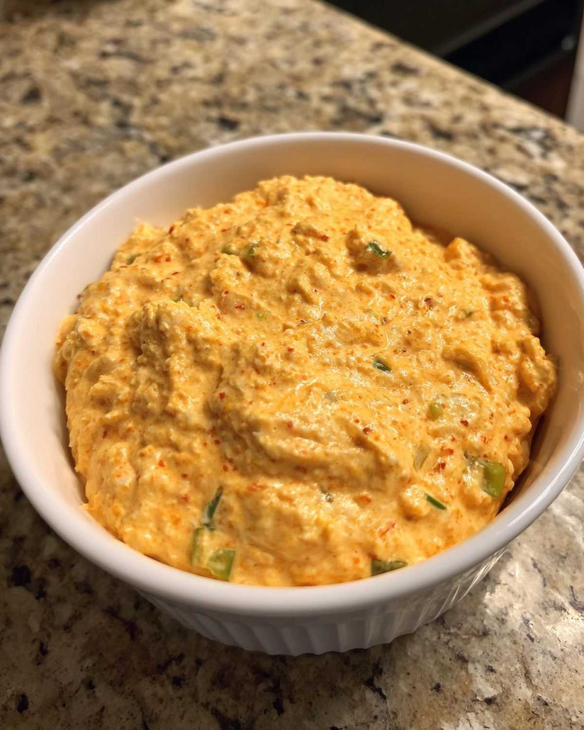 A white bowl filled with creamy, orange-colored cottage cheese queso dip, flecked with green onions and red pepper.
