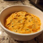 A close-up of bright orange cottage cheese queso dip, flecked with green herbs, served in a white bowl.