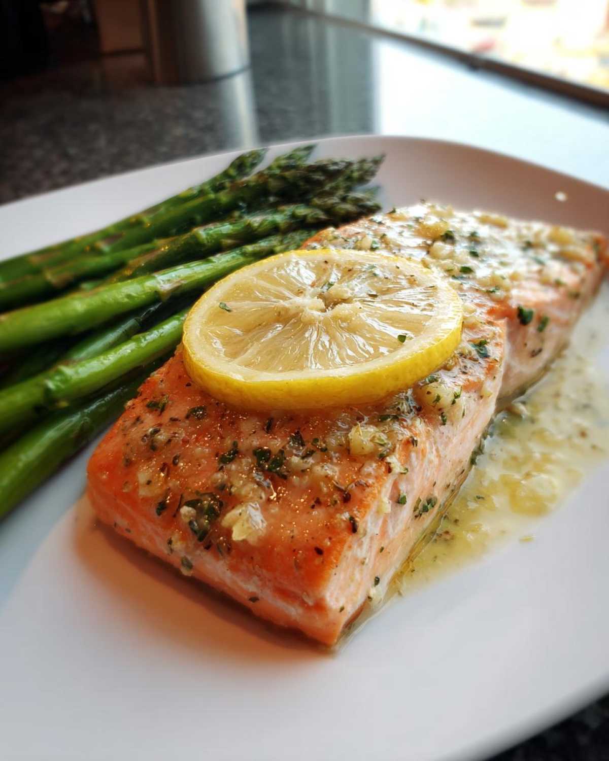 Close-up of a pan-seared salmon recipe fillet topped with a lemon slice, garlic butter sauce, and served with bright green asparagus.