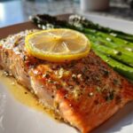 Close-up of a perfectly cooked salmon recipe fillet topped with a lemon slice and herbs, served alongside bright green asparagus.
