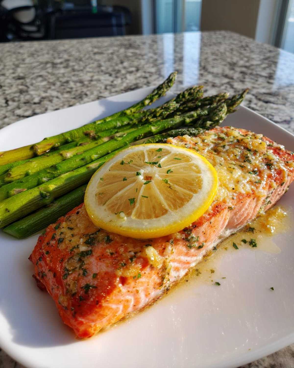 A close-up of a baked salmon recipe fillet topped with lemon butter sauce and a lemon slice, served with roasted asparagus.