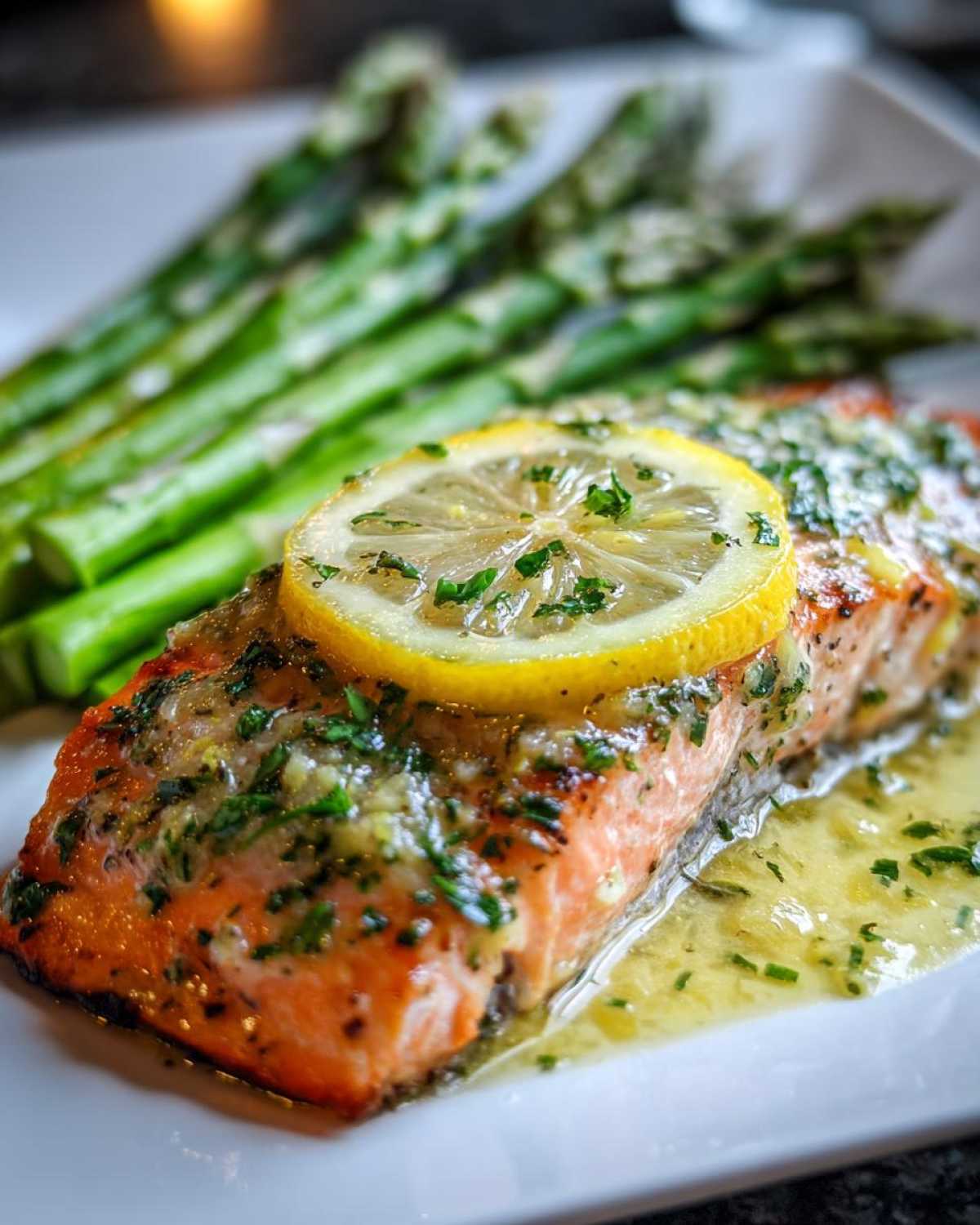 Close-up of a pan-seared salmon recipe fillet topped with lemon and herbs, served alongside bright green asparagus.