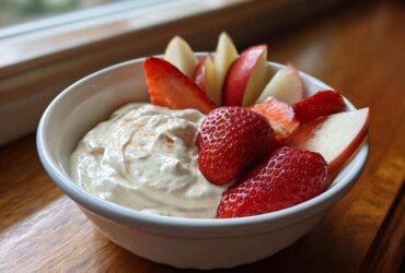 Close-up of a creamy fruit dip in a white bowl served with fresh strawberries and apple slices.