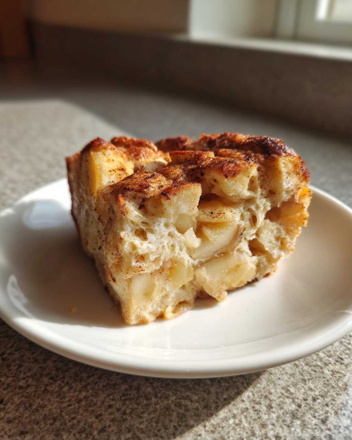 A close-up of a square slice of amazing apple bread pudding, featuring visible chunks of apple and a cinnamon-dusted top.