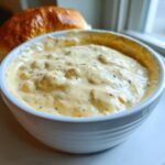 Close-up of creamy, chunky beer cheese dip served in a white bowl next to a golden brown bread roll.