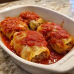 Four perfectly cooked cabbage rolls stuffed with meat, nestled in a white dish and topped generously with bright red tomato sauce.