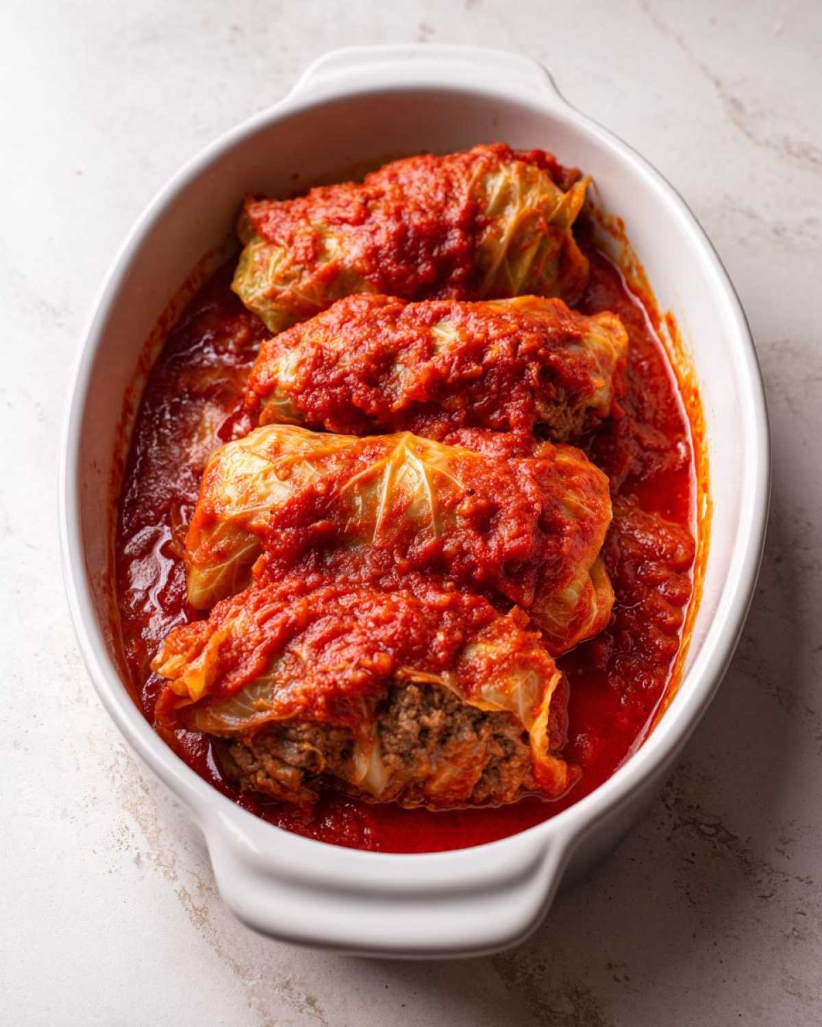 Four hearty cabbage rolls smothered in bright red tomato sauce, resting in a white oval baking dish.