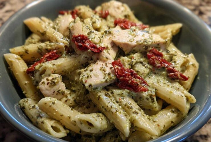 A close-up, overhead view of creamy chicken pesto pasta featuring penne, chunks of white meat chicken, and sun-dried tomatoes in a blue bowl.