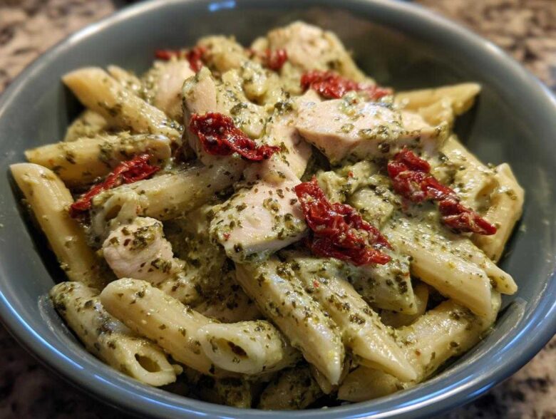 A close-up, overhead view of creamy chicken pesto pasta featuring penne, chunks of white meat chicken, and sun-dried tomatoes in a blue bowl.