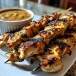 Close-up of perfectly grilled chicken satay skewers with visible char marks, served next to a small bowl of peanut sauce.