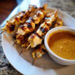 Close-up of grilled chicken satay skewers served on a white plate next to a small bowl of peanut dipping sauce.