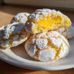 A stack of soft lemon crinkle cookies dusted with powdered sugar, one cookie is broken open showing a bright yellow lemon filling.