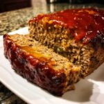 A close-up of a juicy meatloaf, sliced open to show the moist interior and topped with a thick, shiny glaze.
