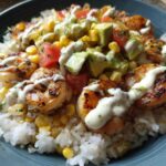 Close-up of a vibrant shrimp bowl featuring grilled shrimp, white rice, corn, avocado, tomatoes, and a creamy drizzle.