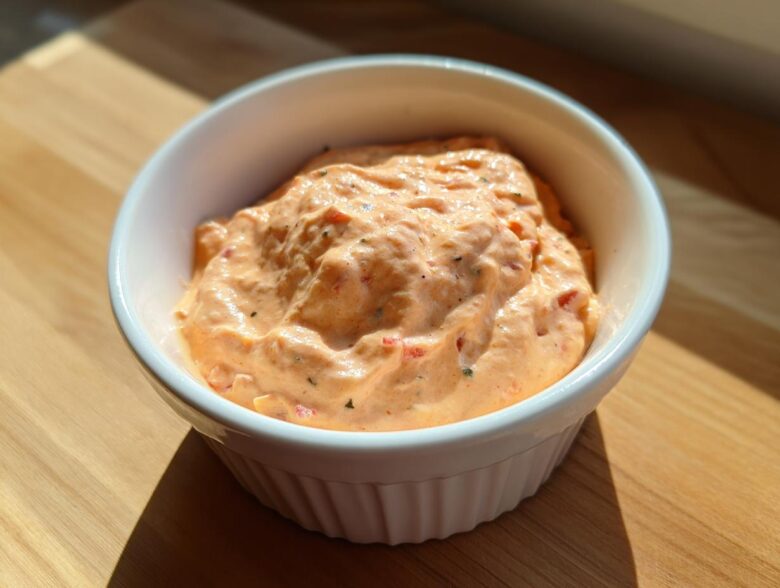 Close-up of creamy, orange-hued smash burger sauce speckled with herbs in a small white bowl.