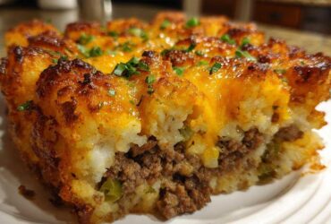 A close-up cross-section of a baked tater tot casserole showing a crispy tater top, melted cheese, and savory ground meat filling.