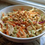 A white bowl filled with vibrant thai chicken salad featuring shredded cabbage, carrots, chicken, and topped generously with chopped peanuts.