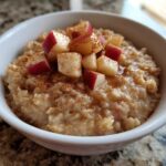 Close-up of a white bowl filled with creamy apple cinnamon oatmeal, topped with diced red apples and a sprinkle of cinnamon.
