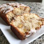 A close-up of a sliced apple fritter bread loaf drizzled with white vanilla glaze on a white plate.