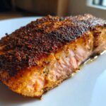 Close-up of a thick, flaky piece of blackened salmon with a dark, spicy crust resting on a white plate.