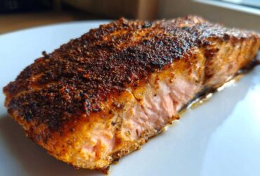 Close-up of a thick, flaky piece of blackened salmon with a dark, spicy crust resting on a white plate.