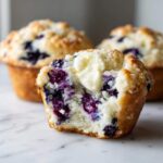 A blueberry cream cheese muffin cut in half showing the moist interior and a dollop of melting butter on top.