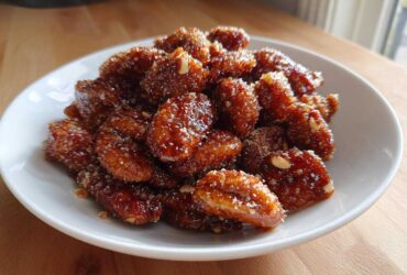 A white bowl filled with glossy, caramelized candied pecans coated in sugar crystals, sitting on a wooden surface.